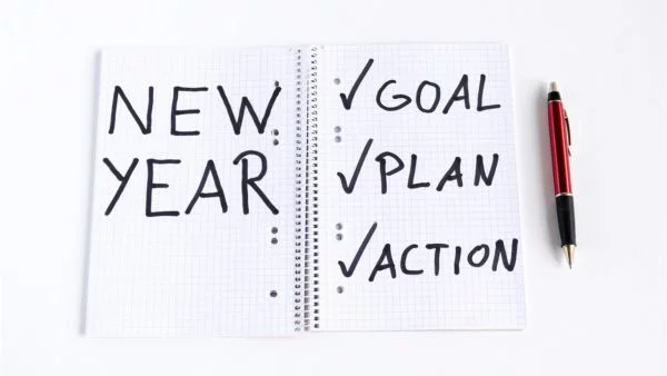 Notebook open to "NEW YEAR" with checkmarks next to "GOAL," "PLAN," and "ACTION." A red pen sits beside it, symbolizing new year resolutions and planning.